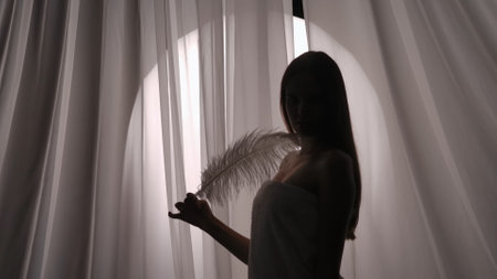 Medium side view shot of a young womans silhouette wrapped in a towel touching and stroking her neck and body with a white feather.の写真素材