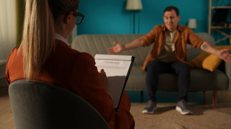 Rear view of a female psychologist against a blurred background of a male patient sitting on a couch in front of her. The therapist is filling out a diagnostic sheet while talking to the male patient.の写真素材