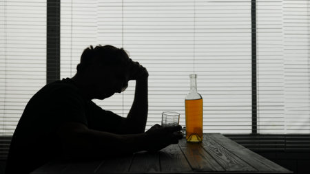 In the silhouette of a young man sitting at a table in a cafe. Nearby is a bottle of alcohol in his hands he holds a glass and thinks about something. He shows alcoholism, sadness and lonelinessの写真素材