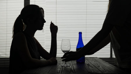 In the frame in silhouette of a young woman she is sitting in a cafe at a table. The waiter has brought her alcohol and a glass. She sits alone and is going to drink. Demonstrates alcoholism, sadnessの写真素材
