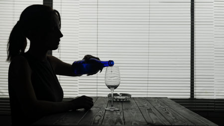In the frame in silhouette of a young woman she is sitting in a cafe at a table. She pours herself alcohol into a glass and is about to drink alone. Demonstrates alcoholism, sadness.の写真素材