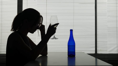 A young woman with glasses is sitting at a table in a cafe thinking about something, a bottle and an ashtray are next to her. She is holding a glass of alcohol. Demonstrates loneliness, alcoholismの写真素材