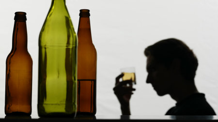 In the close-up shot, there are empty, broken glass bottles full of alcohol. In the background, a strong man holds a glass of alcohol in his hands and examines it. Then he drinks sad and sadの写真素材