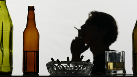 In the close up shot, the camera showing empty, dissipated, glass bottles with alcohol, ash with cigarettes and a glass filled with alcohol. In the background, a man lights a cigarette and smokesの写真素材