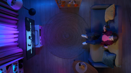 Top view of girl sitting on the couch in the apartment living room, playing video game on TV winning...