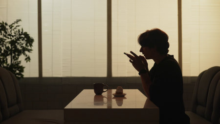 Silhouette of adult male sitting at the bar, man holding smartphone and taking photos of his dessert...