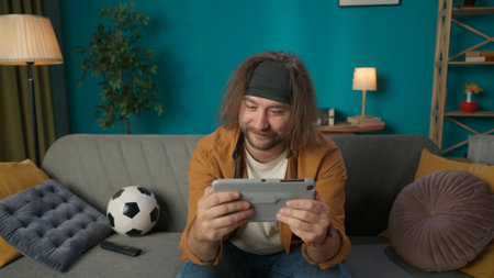 In the shot, a middle aged man is sitting on a sofa in a room. He is holding a tablet and is...