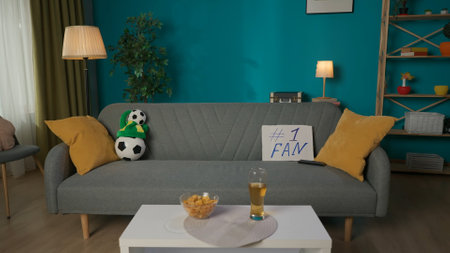 In the frame there is an empty living room with a cozy atmosphere. In the middle of the room there is a gray sofa on which there is a football paraphernalia, a ball, a fan hat and a TV remote controlの写真素材