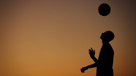 Medium sized photo capturing a silhouette of a teenager, young man playing with a ball outside at sunset. He is throwing the ball in the air.の写真素材