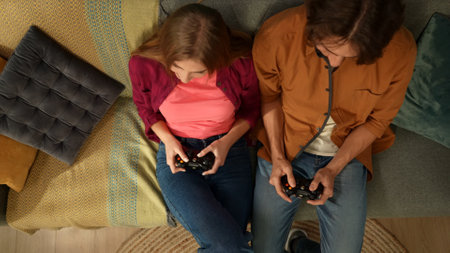Top view capturing a young couple sitting on the couch, holding gamepads, acting as if they are...
