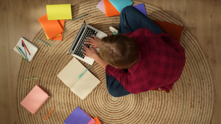 Top view capturing a young woman sitting on the floor and studying, working on a laptop and taking notes, surrounded with colorful books.の写真素材