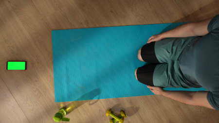Top view capturing an unrecognizable man sitting on a mat in front of a smartphone with an...