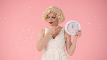 Woman holding a white round wall clock in her hand and pointing her index finger at the time. Woman...