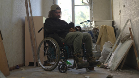 Homeless poor man sitting in a wheelchair in a room of an abandoned building filled with his meager belongings. He is holding a bottle of alcohol, looking frustrated.の写真素材