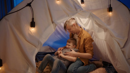 A son lying in the arms of his father in a tent set up in a living room. A man with a little boy sitting on plaids with cushions inside a tent lit with garland.の写真素材