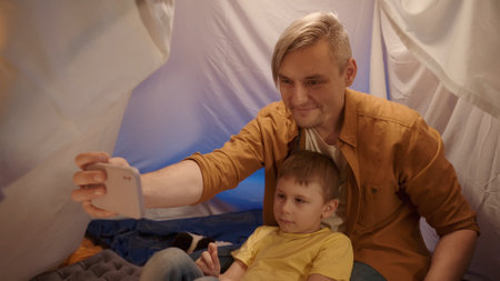 A father and son sit on fur skins in a tent in a dark living room and use a smartphone. Man and boy take selfies together, smiling, close up.の写真素材