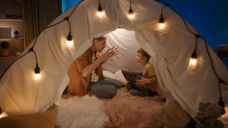 Dad and son spending an evening together. A man and a little boy sit in a tent decorated with holiday lights that stands in a dark living room. Adventures at Home. Concept of happy childhood.の写真素材