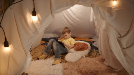 The boy is lying on a large soft toy with a tablet. A little boy spends the evening playing an online game on a tablet, lying in a tent, which is set up in the living room.の写真素材