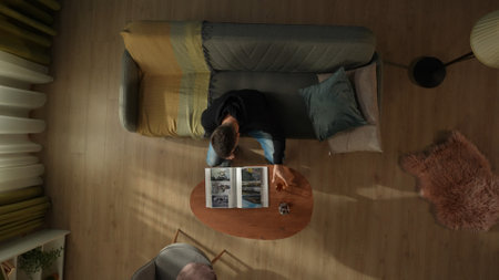 Top view of a man sitting on a living room couch in front of a coffee table with a bottle of alcohol and a glass. Man looking through a photo album. Divorce, separation.の写真素材