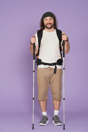 Man tourist in casual clothing with backpack smiling at the camera with trekking poles. Isolated on purple background. Portrait of male traveler.の写真素材