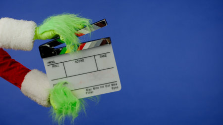 The Grinchs green haired hand holds a movie clapper on an isolated blue background. Gift kidnapper cosplay. Christmas and New Year celebration concept. Blue screen, chroma key.の写真素材
