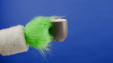 Grinchs green haired hand holds a cup of cocoa with marshmallows on isolated blue background. Gift...