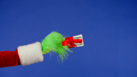 Green haired Grinch hand holds a wad of money wrapped in a red bow on an isolated background. A place for your advertisement. Christmas and new year celebration concept. Blue screen, chroma key.の写真素材