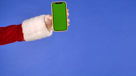 Female hand in Santa Claus costume with green screen phone on isolated blue background. Advertising area, workspace mock up. Christmas and New Year celebration concept.の写真素材