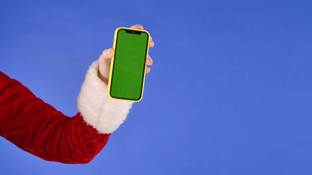 Female hand in Santa Claus costume with green screen phone on isolated blue background. Advertising area, workspace mock up. Christmas and New Year celebration concept.の写真素材