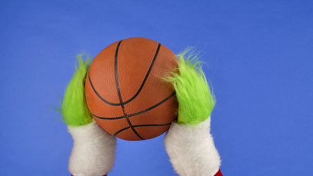 Grinchs green hairy hands holding a basketball on blue isolated background. Gift snatcher cosplay. Christmas and new year celebration concept. Blue screen, chroma key.の写真素材