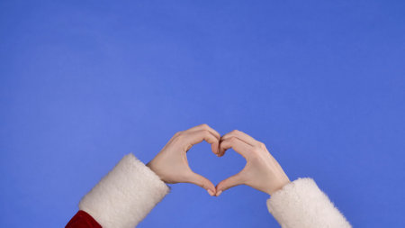 Female hands in Santa Claus costume showing heart sign with fingers on blue isolated background. Christmas and new year celebration concept. Blue screen, chroma key.の写真素材
