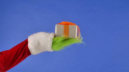 The Grinchs green haired hand holds a white gift box with a orange bow on an isolated blue background. Gift kidnapper cosplay. Blue screen, chroma key.の写真素材