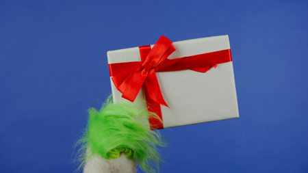 The Grinchs green haired hand holds a white gift box with a red bow on an isolated blue background....