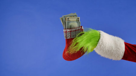 The Grinchs green haired hand hold a Christmas sock with dollar bills on a blue isolated background....