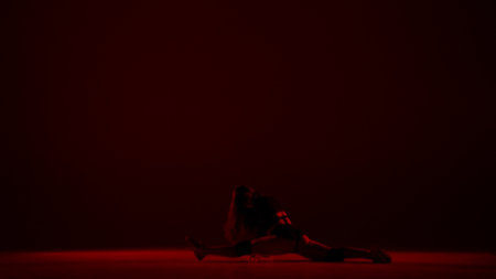 In the frame on a dark background, kneeling woman, a red light shines on her stands a woman. Demonstrates a dance movement in the style of twerk. She is sexy, rhythmic. She is wearing open clothesの写真素材