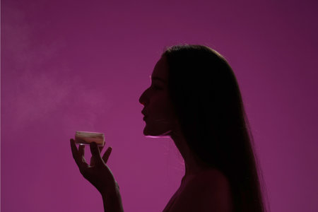 Side view of a dark silhouette of a woman holding a can of powder in a studio on a pink background in pink neon light close up. Seminude woman blowing on powder. Beauty, cosmetology, skin care.の写真素材