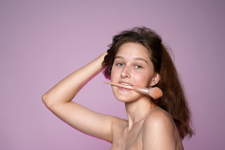 Seminude woman holding a makeup brush in her mouth and holding her long hair with her hand. Portrait of woman in studio on pink background in pink neon light close up.の写真素材