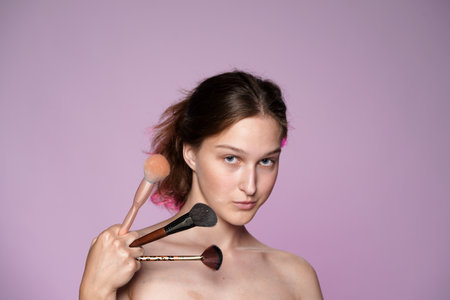 Young woman holding a set of makeup brushes close up. Seminude woman with clear skin and nude makeup in studio on pink background in pink neon light. Face contouring makeup.の写真素材