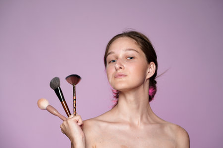 Young woman holding a set of makeup brushes close up. Seminude woman with clear skin and nude makeup in studio on pink background in pink neon light. Face contouring makeup.の写真素材