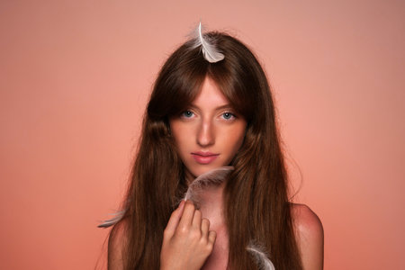 A woman touches the skin of her face with a feather. Portrait of a seminude woman in feathers in studio on orange background close up. Soft and smooth body. Skin care concept.の写真素材