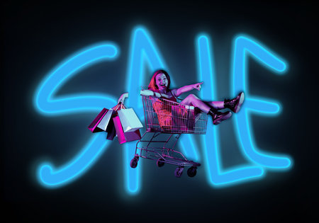 Attractive woman sitting in shopping cart with hands up holding shopping bags, overjoyed face expression. Isolated on background of the word sale.の写真素材