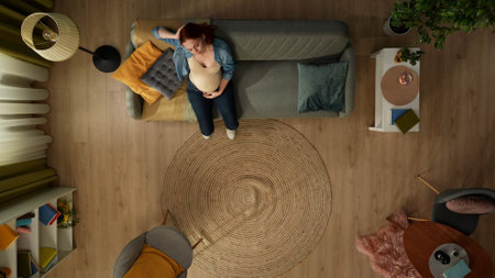 In the shot from above, a pregnant woman sits on a sofa in a room and holds her head. She has a headache, she is irritated, sad. Childrens toys lie on the floor nearby.の写真素材