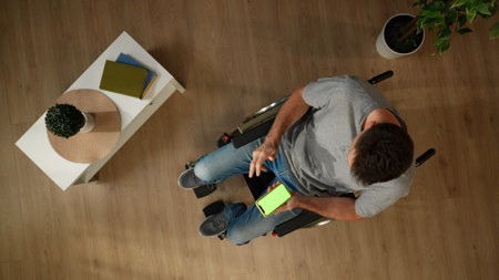 Top view shot a man with mobility impairment sitting on a wheelchair in a room, using a smartphone...