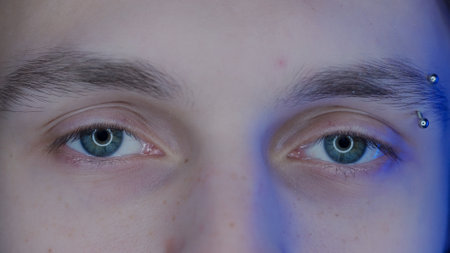 Eyes of young guy with eyebrow piercing macro shot. Blue light from a smartphone, computer screen is...