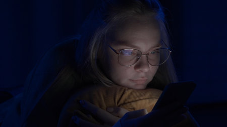 Young woman with glasses is texting on her smartphone lying in bed at home at night close up. Technology, internet, communication and people concept.の写真素材