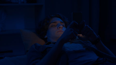 A young man is texting on his smartphone lying in bed at home at night close up. Technology, internet, communication and people concept.の写真素材