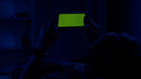 A man lying on the couch at night and holding horizontally a smartphone with a green screen. The man...