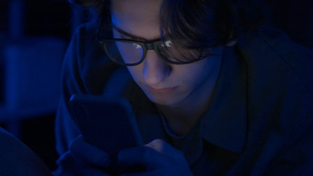 A young man wearing glasses is using a smartphone lying in bed at home at night close up. A man is...
