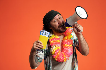 A man speaks into a megaphone and holds up a passport with airline tickets. A man wearing Hawaiian beads in the studio on an orange background close up. Air flight journey concept.の写真素材