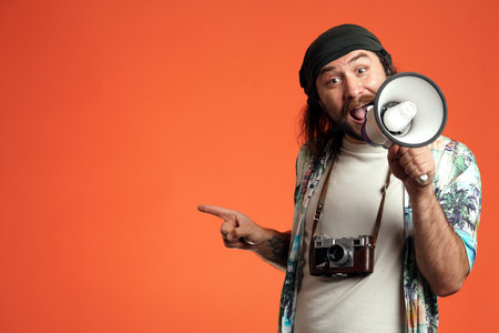 A male traveler with a megaphone points to something with his index finger to the side. A man with a camera around his neck in a studio on an orange background close up. Concept of travel.の写真素材